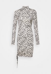 Långärmad, figurnära klänning i kräm med svarta prickar. Har en hög halsringning och sidofållning med en knytdetalj vid fållen.