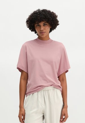 Femme aux cheveux courts et bouclés portant un haut ample rose pâle à manches courtes et un pantalon beige clair à taille élastique sur un fond uni.