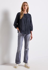Blusa a maniche lunghe blu navy con chiusura a bottoni, yoke strutturato e vestibilità comoda. Abbinata a jeans grigi chiari e sneaker bianche.