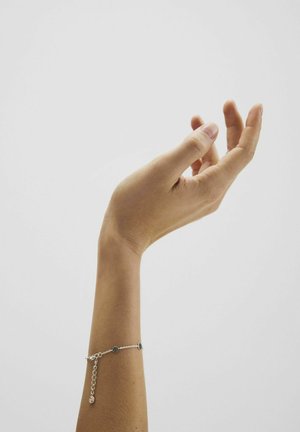 Uniesiona ludzka ręka z lekko zgiętymi palcami, na nadgarstku delikatna srebrna bransoletka z małymi okrągłymi zawieszkami, na jednolitym tle.