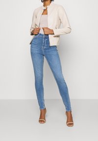 Jasnobeżowa skórzana kurtka z kołnierzem typu stójka, zestawiona z dopasowanymi jasnoniebieskimi jeansami i beżowymi sandałkami na paski. Gładka tekstura, nowoczesny design.