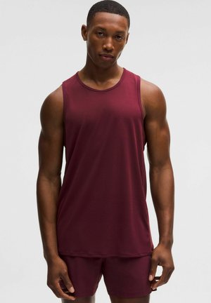 Ung mand iført en ærmeløs bordeaux sports T-shirt og matchende shorts mod en ensfarvet lys baggrund.