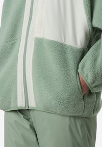 Jachetă verde din fleece cu panouri albe, țesătură texturată, buzunare laterale și închidere cu fermoar. Asortată cu pantaloni de culoare verde deschis.