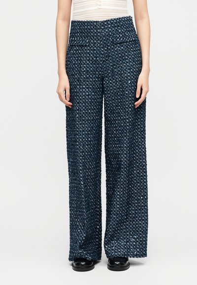 Γυναίκα που φοράει μπλε υφασμάτινο παντελόνι με φαρδιά μπατζάκια και ψηλή μέση, με εμπρόσθιες τσέπες με καπάκι, και μαύρα παπούτσια, στέκεται μπροστά σε απλό φόντο.