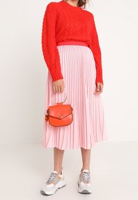 Pull en tricot rouge, jupe plissée rose, sac à main orange avec une anse en anneau, et baskets blanches sportives avec des accents gris et beige.