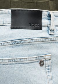 Ανοιχτό μπλε τζιν υφασμάτινο παντελόνι με μαύρο δερμάτινο έμπλαστρο με ανάγλυφη την επιγραφή «BOSS» και ένα μικρό μεταλλικό λογότυπο «B» κοντά στην πίσω τσέπη.