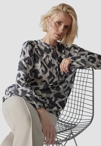 Bluse in Hellgrau mit schwarzem und cremefarbenem abstraktem Muster, mit Rundhalsausschnitt und langen Ärmeln mit elastischen Bündchen, kombiniert mit beigen Hosen.