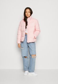 Lys rosa pufferjakke med høy krage og glidelås foran, med en logo på brystet. Brukt med slitte lyseblå jeans og hvite joggesko.