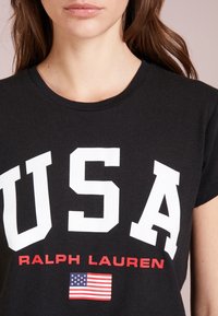 Černé bavlněné tričko s velkým bílým textem "USA", červeným "RALPH LAUREN" a malou grafikou americké vlajky pod ním. Krátké rukávy.