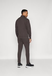 Dunkelbrauner Trainingsanzug mit hohem Kragen und figurbetontem Design. Hergestellt aus weichem Material, mit gerippten Bündchen und Seitentaschen. Schwarze Turnschuhe.