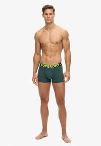 Bóxers verdes con una amplia cinturilla negra que presenta 'SUPERDRY' en amarillo brillante. Tejido suave con un corte ajustado. Sin patrones visibles.