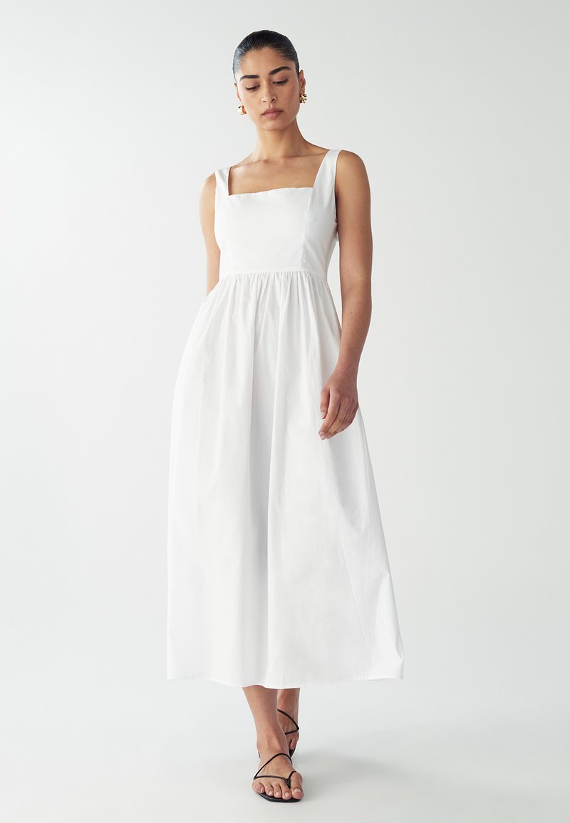 Valkoinen, hihaton mekko, jossa on neliömäinen pääntie, kerätty vyötärö ja lattian pituinen hame. Silkkinen puuvillakangas, jossa on puhdas ja minimalistinen muotoilu.
