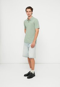 Šviesiai žalias polo marškinėlis su subtiliu logotipu, derinamas su šviesiai mėlynais džinsiniais šortais. Juodi slip-on batai ir kreminės spalvos kojinės užbaigia aprangą.