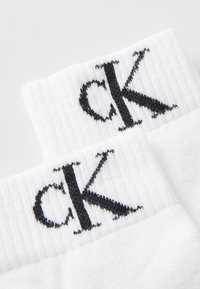 Biele bavlnené ponožky s krátkym strihom a rebrovanou textúrou; na manžetách je výrazne vyšité čierne logo "ck".