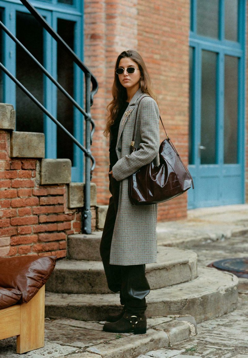 Šedý houndstooth dlouhý kabát, černý top, kožené kalhoty, hnědá kabelka, kulaté černé sluneční brýle a tmavé boty, na pozadí cihel a modrých oken.