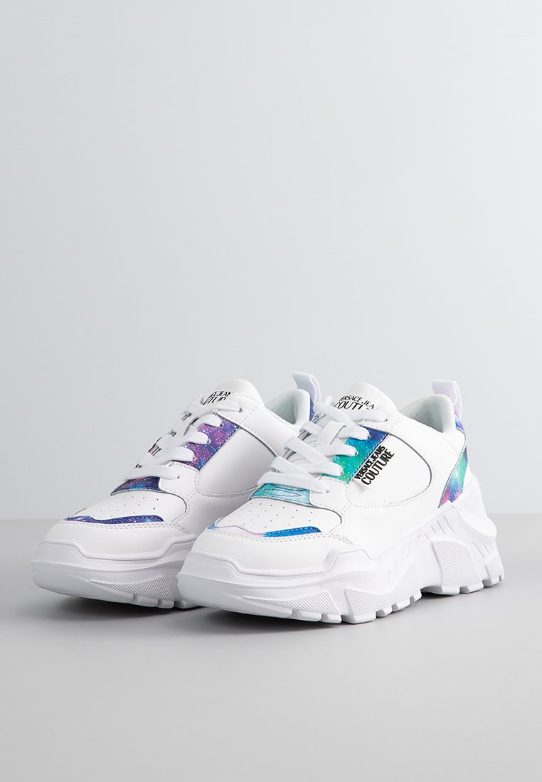 Zapatillas gruesas blancas con acentos iridiscentes multicolores y la marca "Versace Jeans Couture" en negro en el lateral y la lengüeta, sobre una superficie gris.