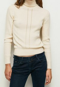 Kremowy sweter z golfem o żebrowanym wzorze z pionowymi teksturami i akcentującymi otworami. Dopasowany krój, długie rękawy i żebrowany dół.