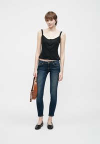 Top corto negro con botones, combinado con jeans ajustados azul oscuro. La modelo sostiene un bolso marrón de hombro y lleva plano negro. Fondo claro.