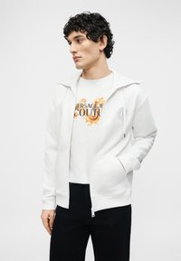 Νέος άνδρας που φοράει μια λευκή ζακέτα με φερμουάρ πάνω από λευκό T-shirt με σχέδιο και μαύρο παντελόνι, στέκεται με το ένα χέρι στην τσέπη.