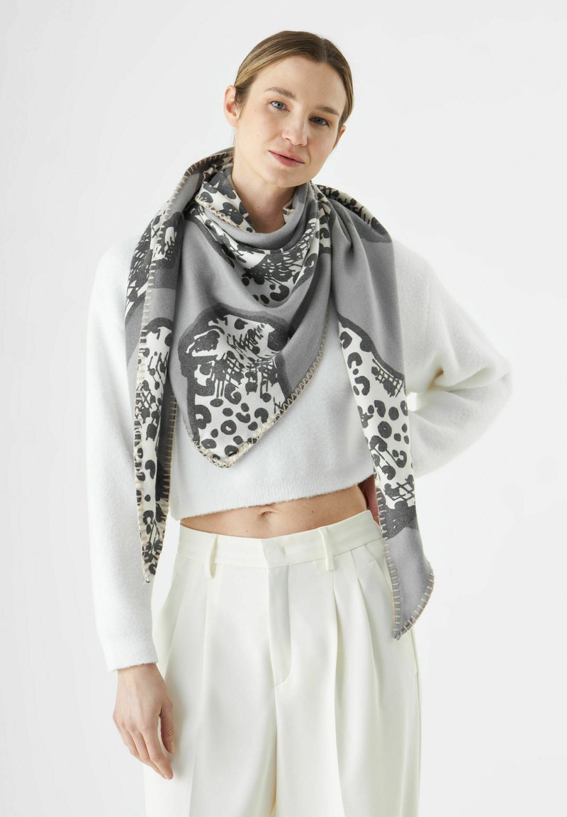 Écharpe grise et blanche à motifs léopard et motifs animaliers, de forme triangulaire et texture douce, portée sur un sweat crop blanc.