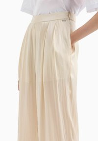 Pantaloni beige plissettati con una texture liscia e lucida e tasche laterali. Caratterizzati da una vita alta e un piccolo logo in metallo.