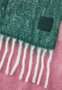 Grøn og pink uld tørklæde med en tekstureret overflade, der har hvide frynser og et sort logo-patch. Blød og koselig design.