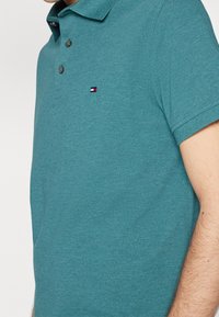 Tyrkysové polo tričko vyrobené z texturované látky. Má límeček, tři knoflíky a malou nášivku v červené, bílé a modré barvě na prsou.