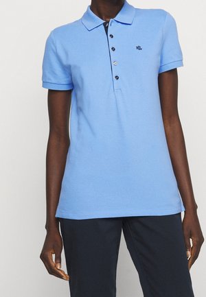 Lyseblå polo med korte ærmer, krave og fem sorte knapper, båret sammen med mørke bukser på en person, der vises fra skuldre til hofter.