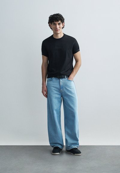 Μαύρο κοντομάνικο μπλουζάκι με ανάγλυφο λογότυπο, συνδυασμένο με τζιν παντελόνι σε ανοιχτό μπλε χρώμα με φαρδιά μπατζάκια και μαύρα αθλητικά παπούτσια. Απλό φόντο.