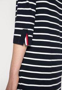 Haut à rayures bleu marine et blanches avec manches courtes, doté d'une fente latérale agrémentée de rayures rouges et blanches à l'ouverture. Texture en tricot doux.