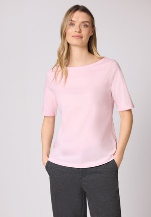 Femme portant un haut à manches courtes rose clair et un pantalon gris foncé, debout les mains dans les poches devant un fond uni.