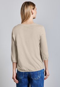 Beige, leichtes Langarmoberteil mit rundem Ausschnitt und gerafften Ärmelbündchen, kombiniert mit blauen Denim-Jeans.