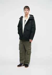 Čierna kapucňa s zipsom zo prednej strany, bočné vrecká a nastaviteľné manžety, skombinované s olivovými cargo nohavicami a čiernymi teniskami.