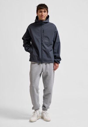 Donkergrijze waterdichte jas met een capuchon en een voorzak met rits, gecombineerd met lichtgrijze joggingsbroek en witte sneakers. Eenvoudig, functioneel ontwerp.