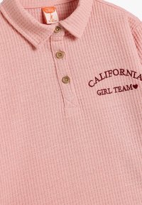 Lyserød ribbet polo-shirt med krave, tre brune knapper og "CALIFORNIA GIRL TEAM" broderet i mørk burgunder.