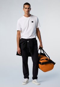 Maglietta bianca con un piccolo logo nero, pantaloni neri, sneaker bianche e una borsa a tracolla arancione con accenti e tracolla neri.