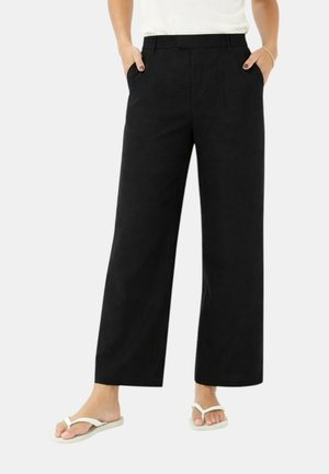 Donna che indossa pantaloni neri a gamba larga con le mani nelle tasche, abbinati a sandali infradito bianchi su uno sfondo semplice.