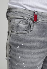 Gros plan sur un jean en denim gris avec des éclaboussures de peinture blanche, porté avec une chemise blanche, présentant une étiquette de marque rouge sur la passants de ceinture.