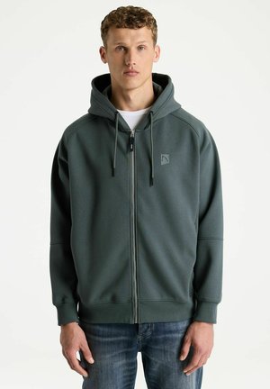 Young man with curly hair wearing dark green zip-up hoodie and blue jeans standing against white background.