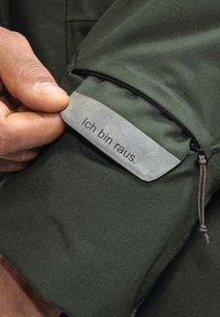 Dunkelgrüner Jackenstoff mit sichtbarer Tasche, ausgestattet mit einem silbernen Etikett mit geprägt Text "Ich bin raus." Reißverschluss und Kordelzugdetails sind vorhanden.