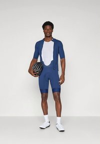 Fato de ciclismo azul-marinho com mangas curtas e painéis em rede, apresentando calções acolchoados. Modelo segura um capacete preto, calçando sapatos e meias brancos.