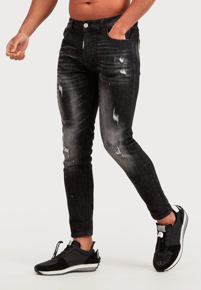 Homme portant un jean skinny noir usé et des baskets noires, debout avec une main dans la poche devant un fond uni.