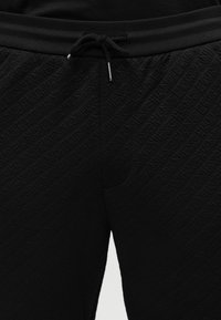 Μαύροι παντελόνια με υφή, ελαστική μέση και μαύρο κορδόνι με διακριτικό ανάγλυφο διαγώνιο μοτίβο κειμένου.