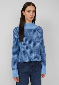 Vrouw met lang, donker haar, gekleed in een blauw, structuurgebreid sweater met een geribbelde hoge kraag en manchetten, in combinatie met donkerblauwe jeans, staande tegen een grijze muur.