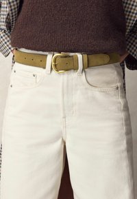 Cintura in camoscio verde con una fibbia in metallo dorato, indossata sopra jeans bianchi in denim con due tasche frontali e dettagli di cucitura. Top in maglia strutturata visibile.