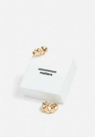 Gold twisted hoop earrings beside a white square box labeled “uncommon matters” in bold black text. Smooth metallic finish, sculptural design.