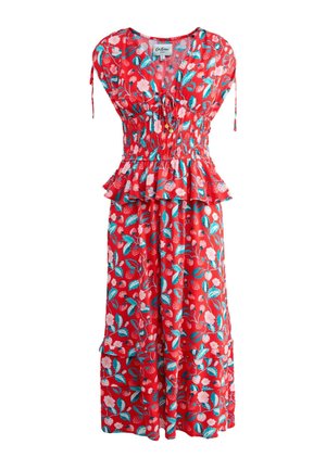 Frau steht vor einer pinkfarbenen Wand, trägt ein rotes ärmelloses Kleid mit Blumenmuster und Rüschensaum an der Taille, neben einem weißen Stuhl mit Kissen und kariertem Boden.