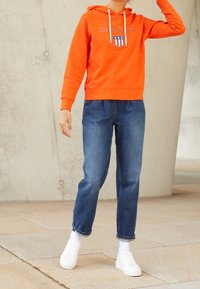 Pomarańczowa bluza z kapturem z logo i białymi sznurkami, zestawiona z niebieskimi jeansami i białymi trampkami, na tle betonowej ściany.
