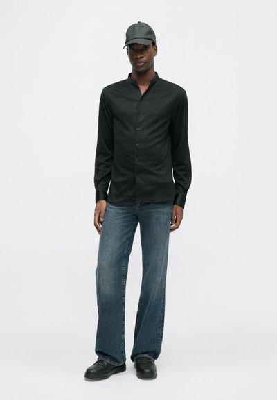 Mies, joka pitää päällään mustaa napillista paitaa, mustaa lippistä, sinisiä farkkuja ja mustia kenkiä, seisoo yksinkertaista valkoista taustaa vasten.