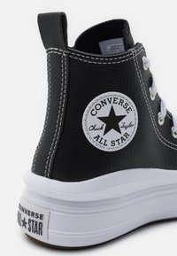 Sneaker alta in ecopelle nera con cuciture e lacci bianchi. Presenta una toppa rotonda con un logo a stella e una suola in gomma testurizzata.